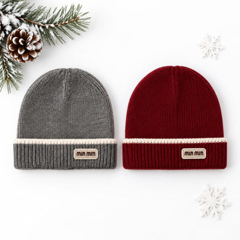 Two ribbed knit beanie hats laid flat on a white background: one heather gray and one deep red, each with a cream stripe and a small tan 'min min' patch on the cuff. Pine branches and snowflakes are styled around them.