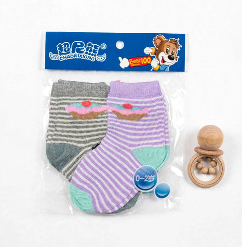 A pair of baby socks in a clear plastic bag with a blue branded header card. One sock is gray and white striped with a pink cupcake graphic, and the other is lavender and white striped with a mint green heel/toe area and a pink cupcake graphic.