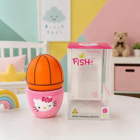 A pink and orange fish-shaped baby feeding bottle with a plush Hello Kitty base, shown next to its clear retail box on a white tabletop. The background is a pastel-colored children's room with a crib, wall art, and a teddy bear.